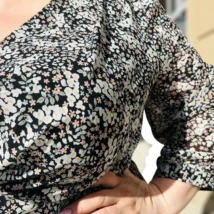 Lili sukienka asymetryczna: detal dekoltu i rękawa 3/4, widoczny drobny, kwiatowy wzór na czarnym tle.